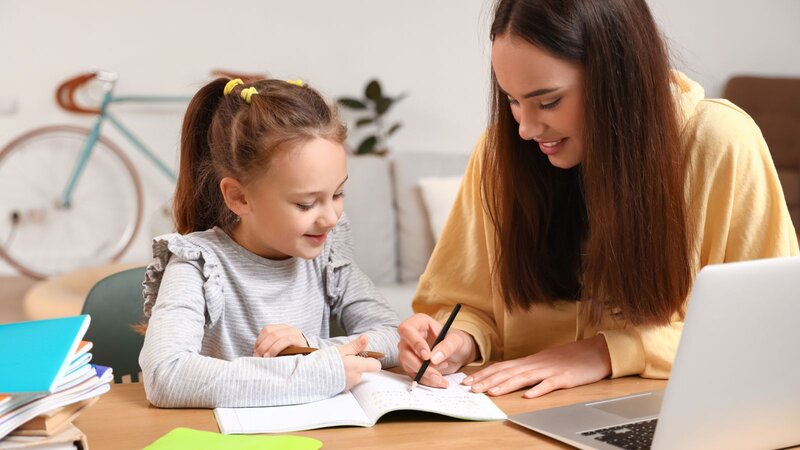 Woman helping a young girl with schoolwork at a table.