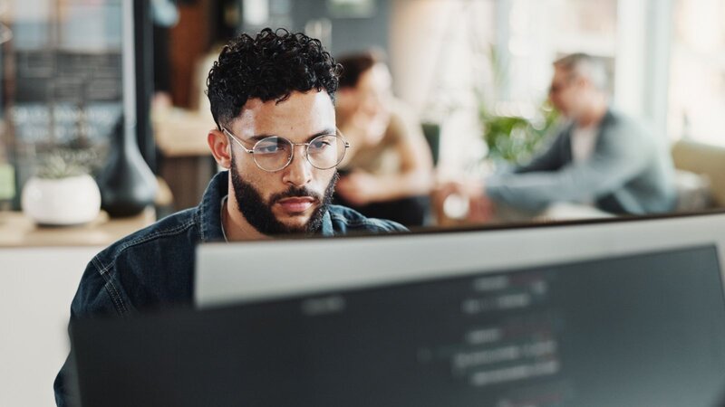 A man with curly hair and glasses focused on a computer screen in a modern office setting.