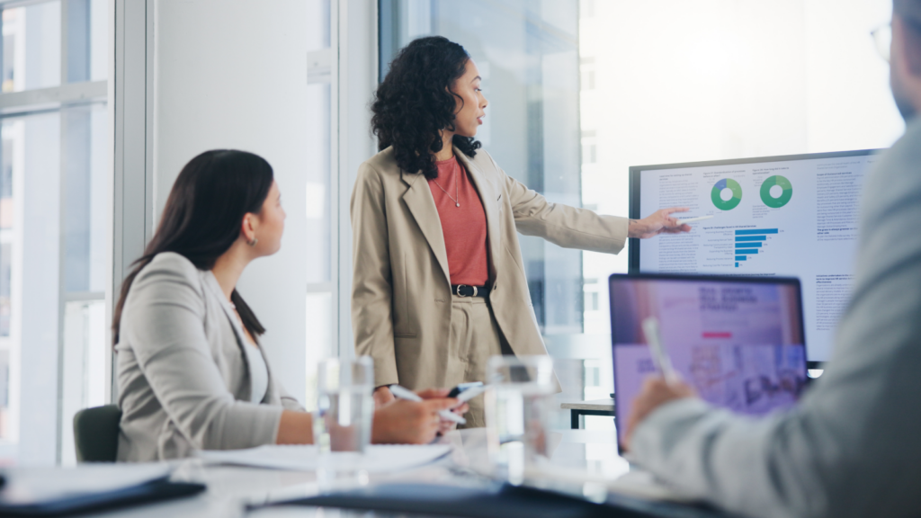 A woman stands at a podium, presenting to her colleagues seated at a conference table, engaged in the discussion.