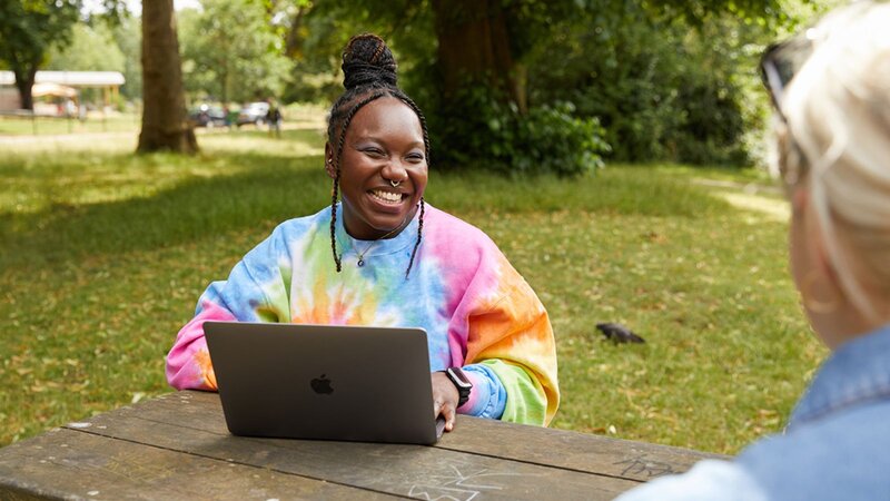 A woman wearing a colorful tie-dye sweater sitting at a picnic table with a laptop, smiling at another person. Outdoor greenery in the background.