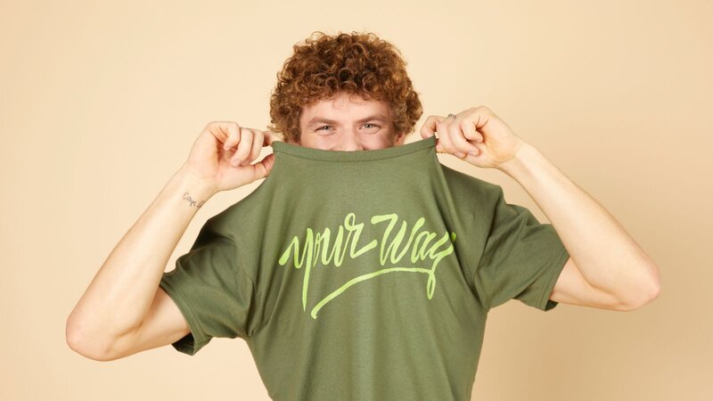 A man with curly hair playfully pulling up a green t-shirt with "Your Way" written in yellow cursive on it, against a light-colored background.