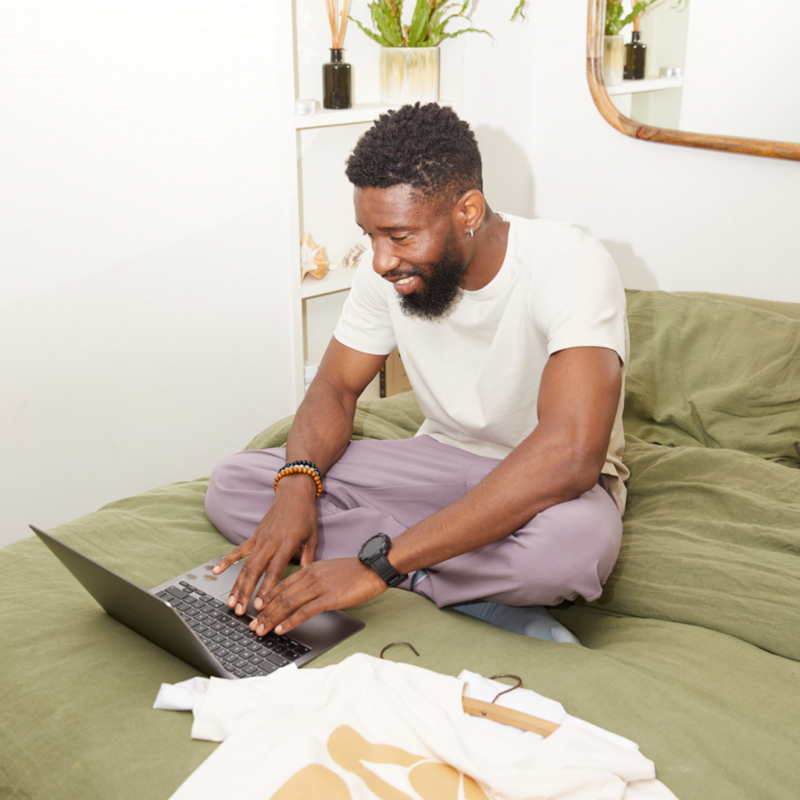 Man sitting cross-legged on a bed with green sheets, smiling while using a laptop. He's wearing a white t-shirt and purple pants.