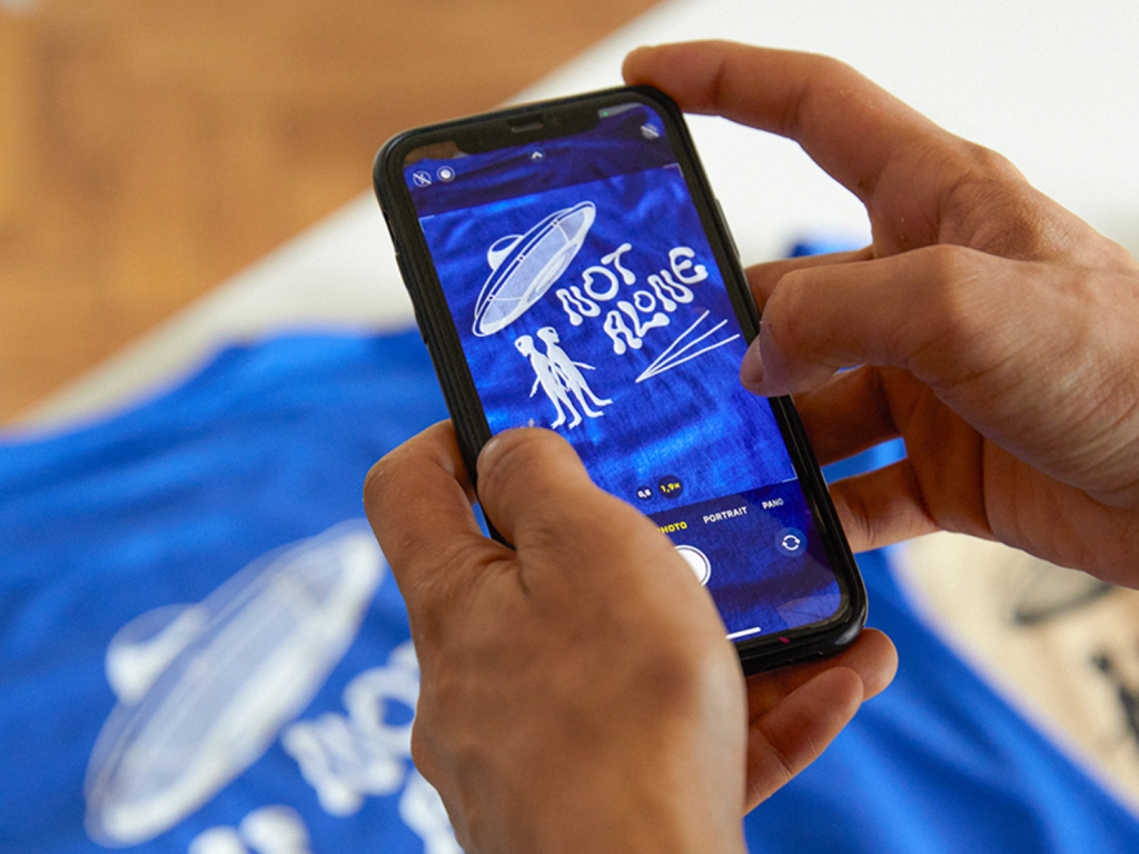 A person taking a picture of a blue t-shirt with an alien-themed design and “Not alone” text print.