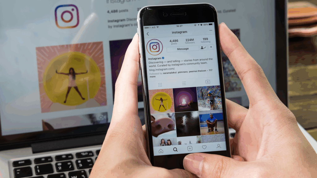 A person holding a mobile phone with the Instagram app on the screen, with a blurred laptop in the background.