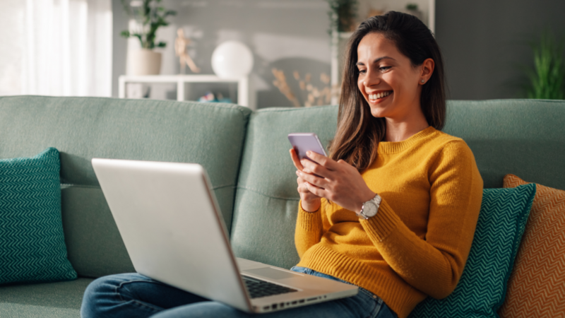 A smiling woman in a yellow sweater sits on a teal sofa, holding a smartphone in front of a laptop.