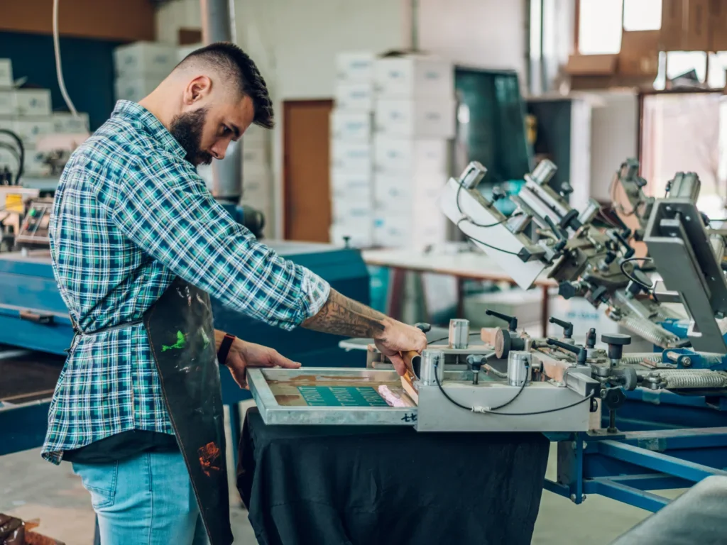 A man operates a screen printing machine, focused on creating custom t-shirts.
