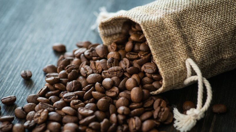 Roasted coffee beans spill out of a small burlap drawstring bag onto a dark wooden surface.