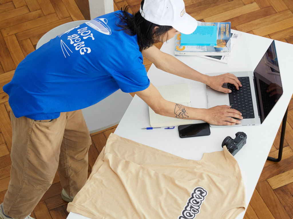 A person typing on a laptop keyboard that sits on top of a table with a custom t-shirt near it.