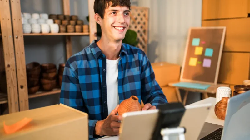 A smiling man holding a ceramic vase, sitting at a cluttered work desk with a laptop and packages. Pottery and a board with notes behind him.