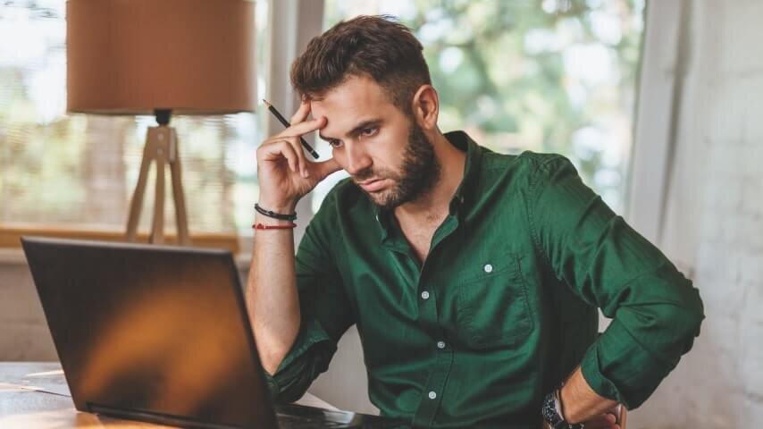 A man in a green shirt looking at a laptop screen and thinking.