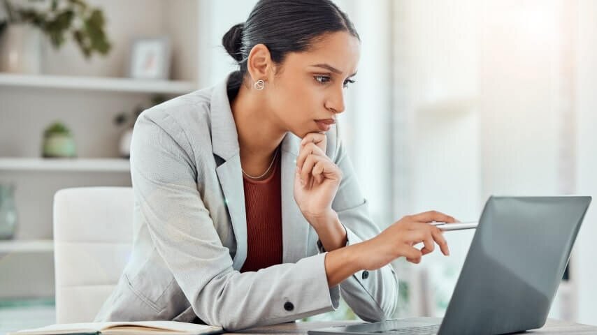 A woman in a white blazer looking at a laptop screen, pointing with a pen.