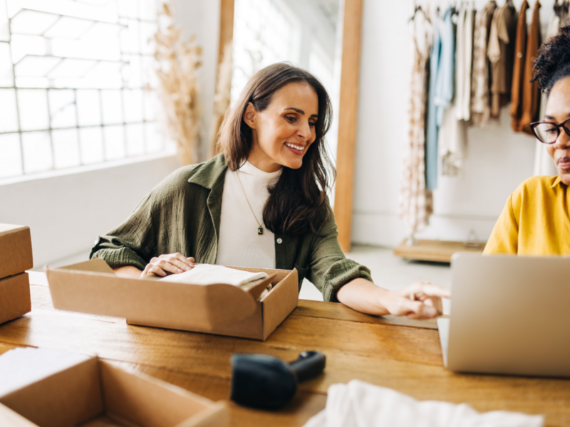 A woman preparing a delivery box for shipment while conversing with another woman.