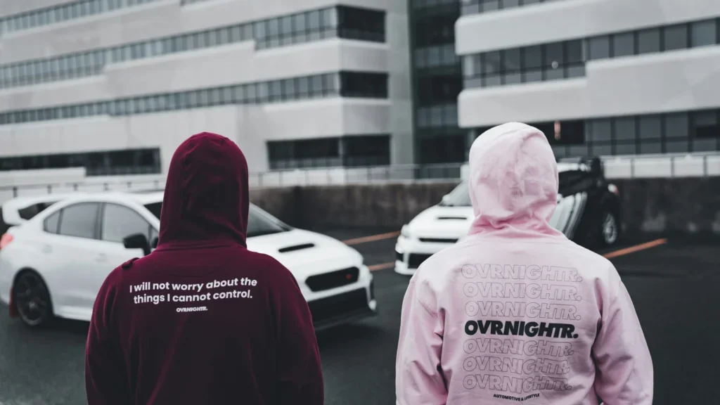 Two men facing two parked cars wearing custom burgundy and pink hoodies with a back print.