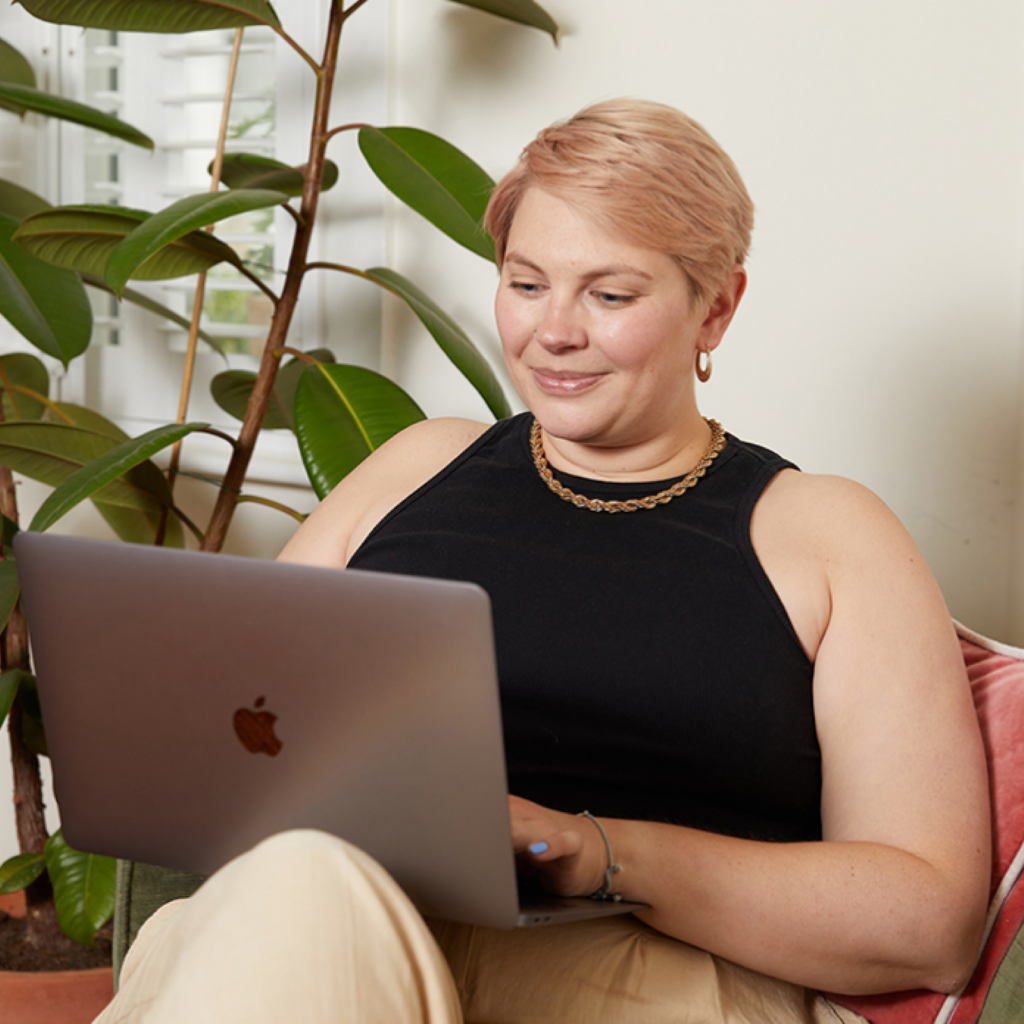 A woman with short hair sits on a sofa, smiling at a laptop in a cozy setting. A large green plant is nearby.