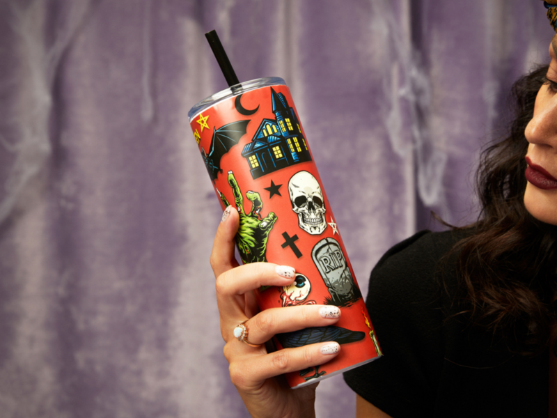 A woman holding a Halloween-themed cup, featuring spooky designs and vibrant colors, smiles in a festive atmosphere.