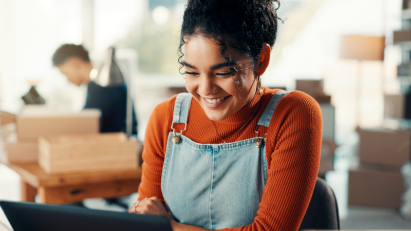 A smiling woman in an orange sweater and jeans overalls is looking at a tablet.