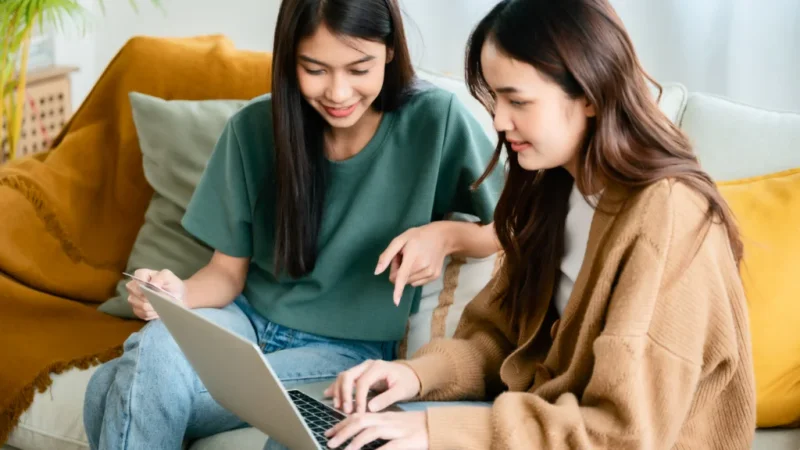 Two women are sitting on a yellow couch, shopping online, with one woman holding a laptop and the other a debit card.