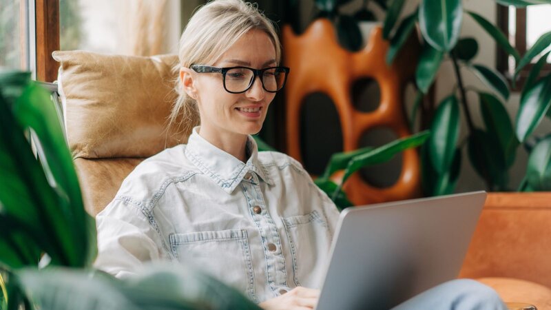 A woman sitting on a couch, smiling while working on a laptop surrounded by potted plants.