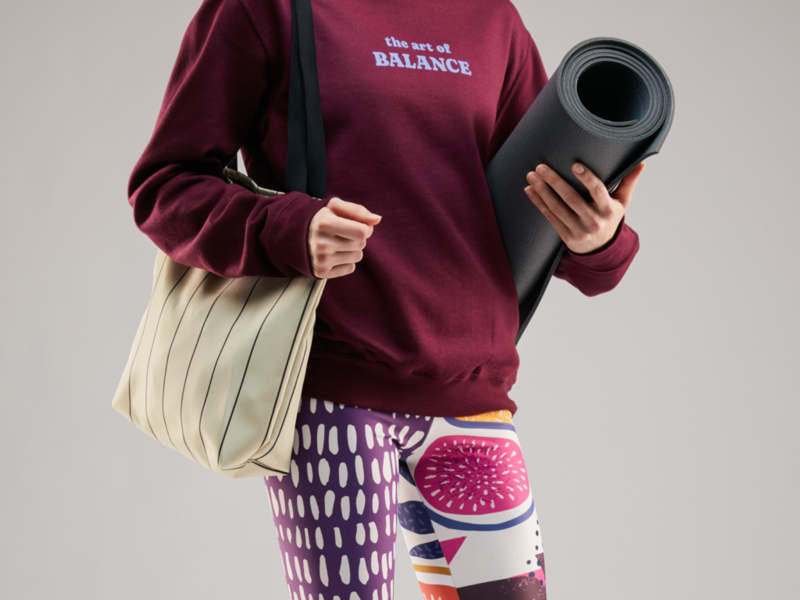A mockup of a woman in a custom hoodie and yoga pants, holding to a yoga mat and a tote bag.
