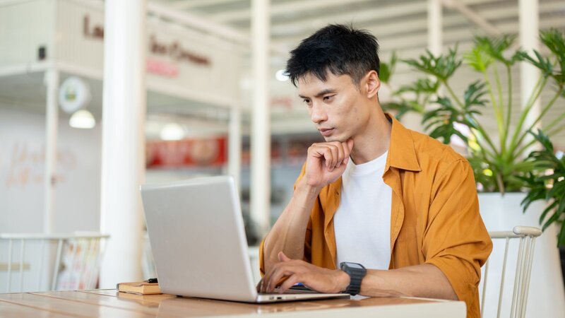 A young man in an orange shirt sits at a table, working intently on a laptop in a spacious, well-lit indoor setting with plants in the background.