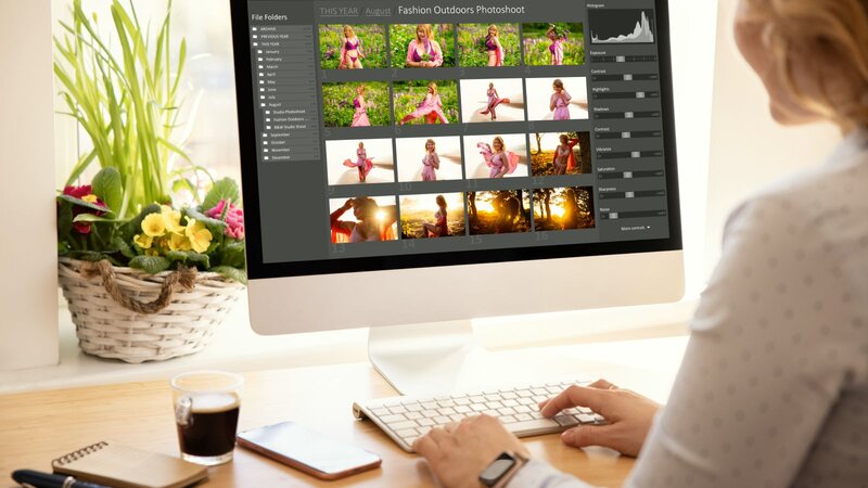 A person editing photos on a computer at a desk with a coffee cup and a flower basket in the background.