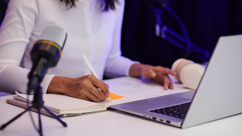 A person writing in a notebook while using a laptop, with microphones in the background.