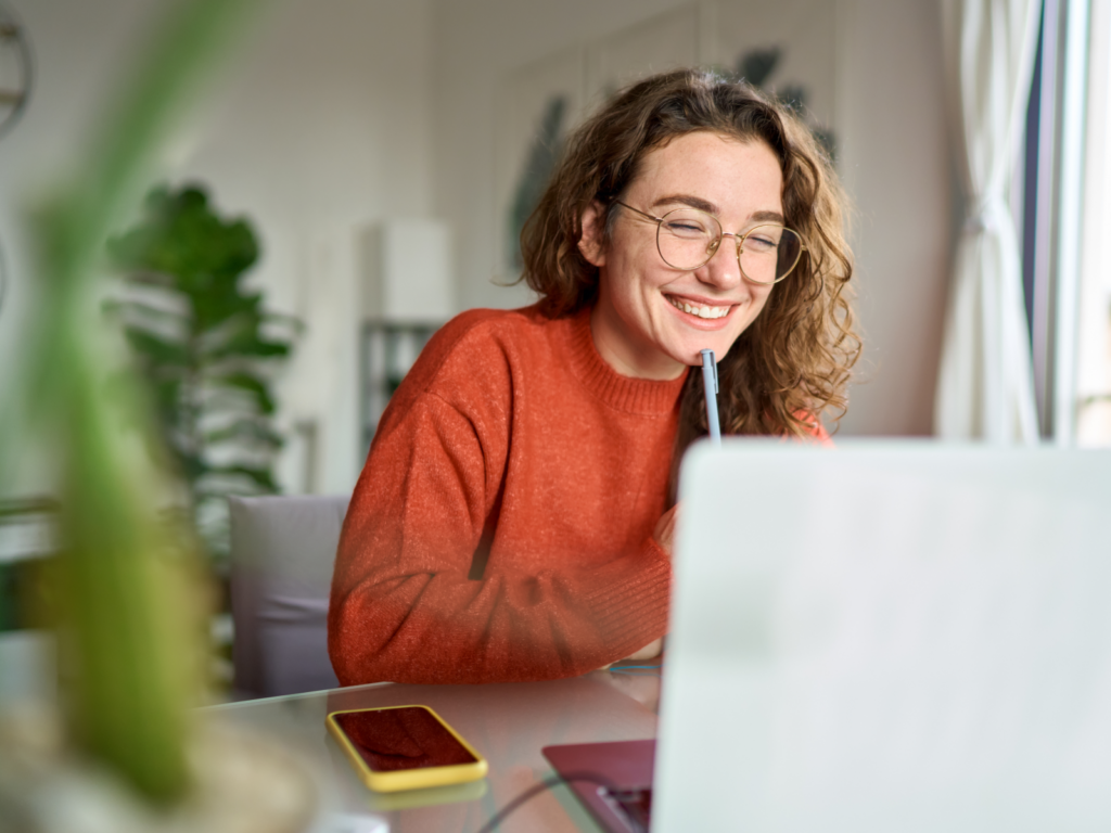 A smiling woman with glasses happily uses her laptop, radiating positivity and focus.