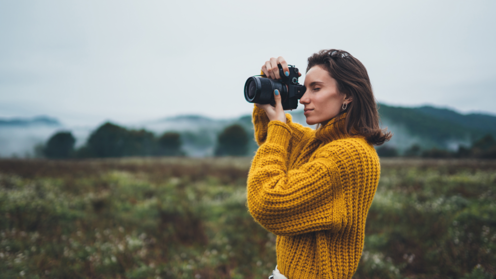 A woman in a yellow sweater captures a moment with her camera in nature.