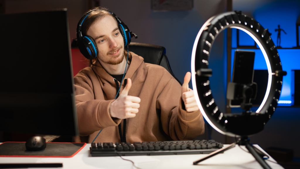 A man wearing headphones sits at a computer, smiling and giving a thumbs up for a video game.