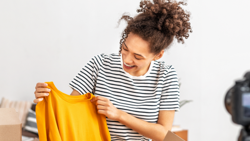 A woman in a white and black-striped shirt is holding a yellow t-shirt, recording the process of unboxing using a camera.