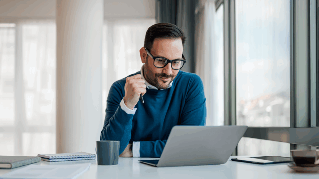 A man wearing glasses is seated at a desk, working on a laptop.
