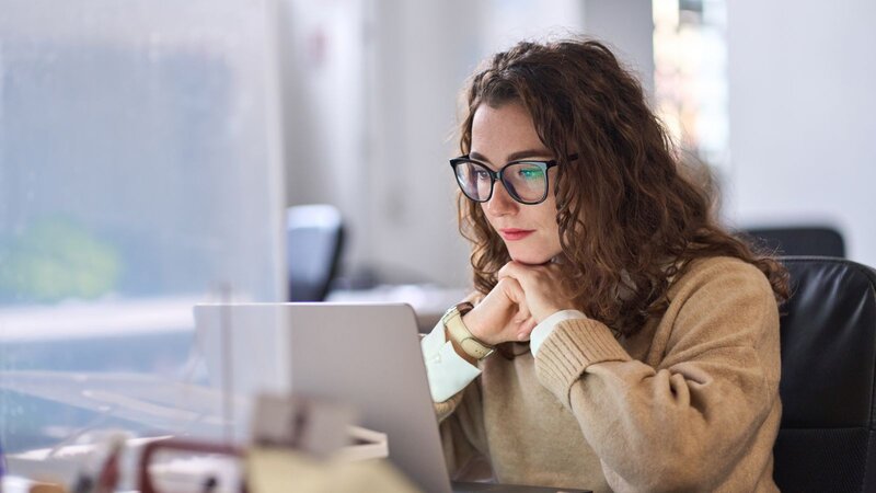 A woman with curly hair and glasses sitting at a desk, focused on her laptop, learning how to sell digital planners on Etsy.