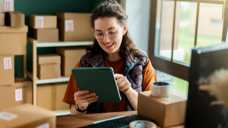 A woman is sitting at a desk in a room filled with cardboard boxes, smiling as she looks at a tablet. She is wearing glasses and a vest.