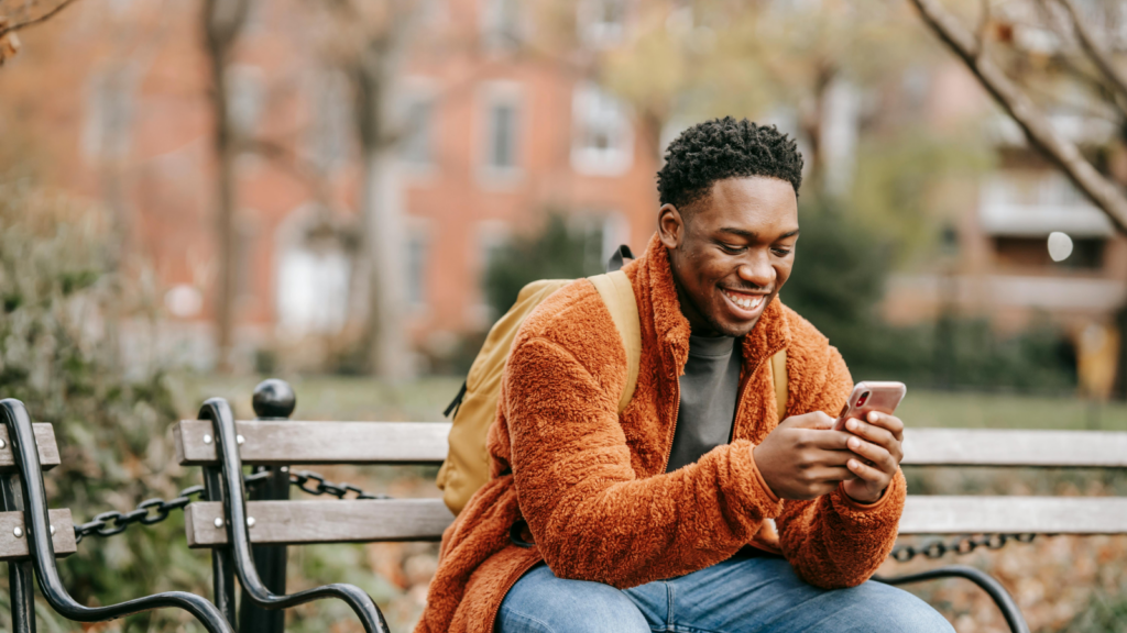 A smiling man sitting on a park bench and using a mobile phone he’s holding in his hands.