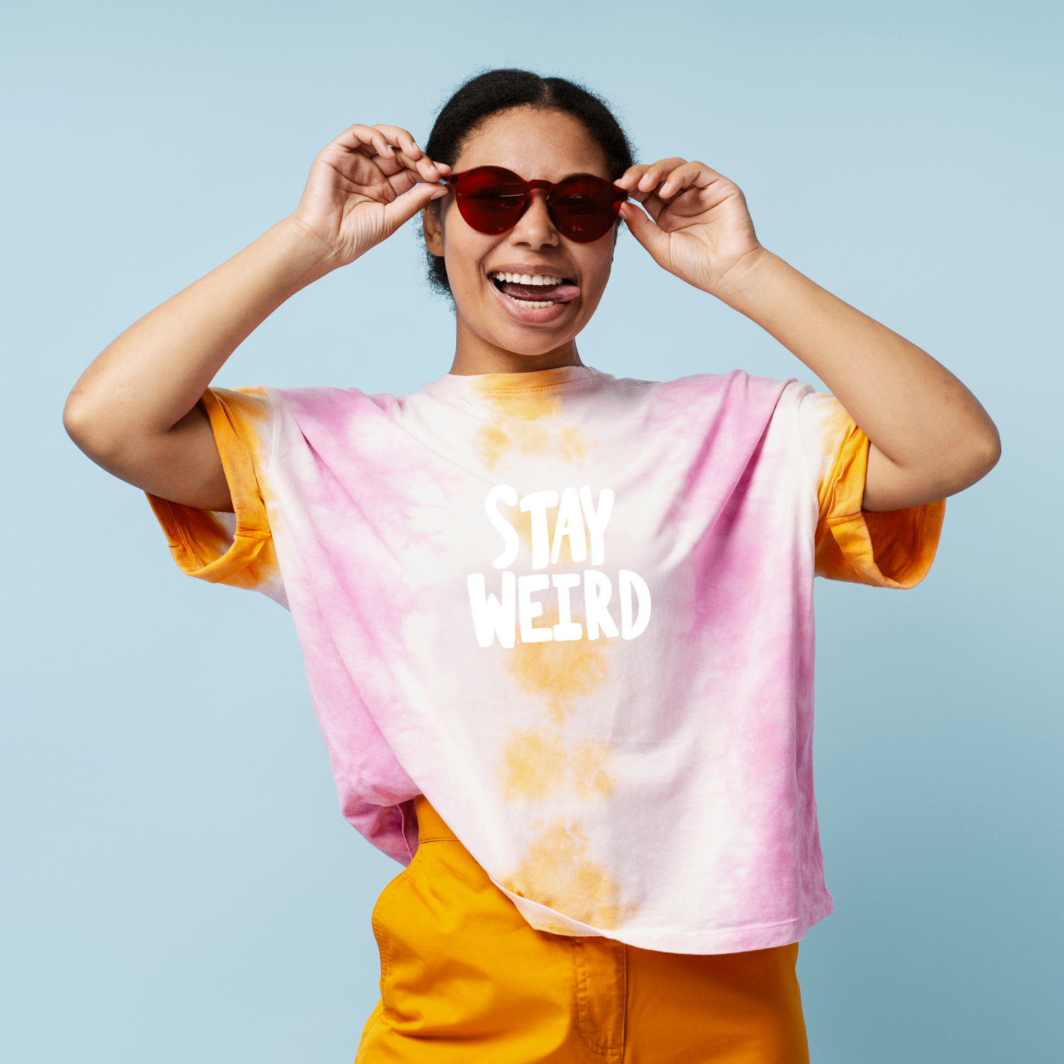 Smiling person in sunglasses and a colorful tie-dye shirt with "Stay Weird" text, posing playfully against a light blue background.
