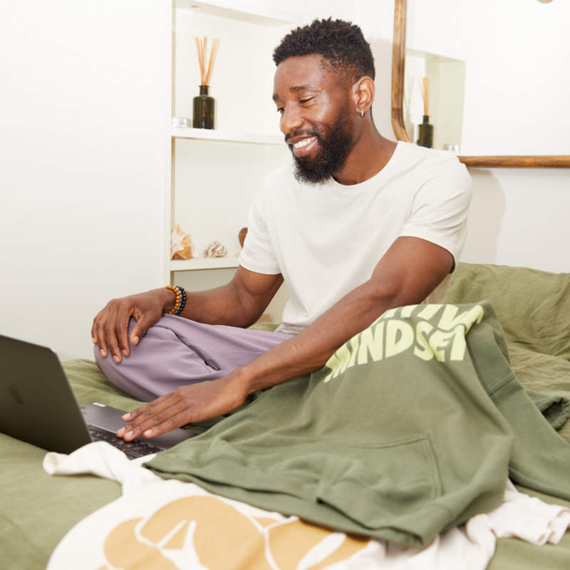 A smiling man sitting on a couch with a laptop and a custom “Positive mindset” hoodie.