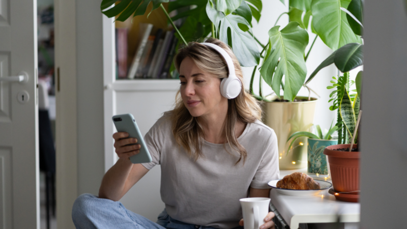 A woman is sitting in a kitchen, wearing headphones, drinking from a mug, and scrolling through her phone.