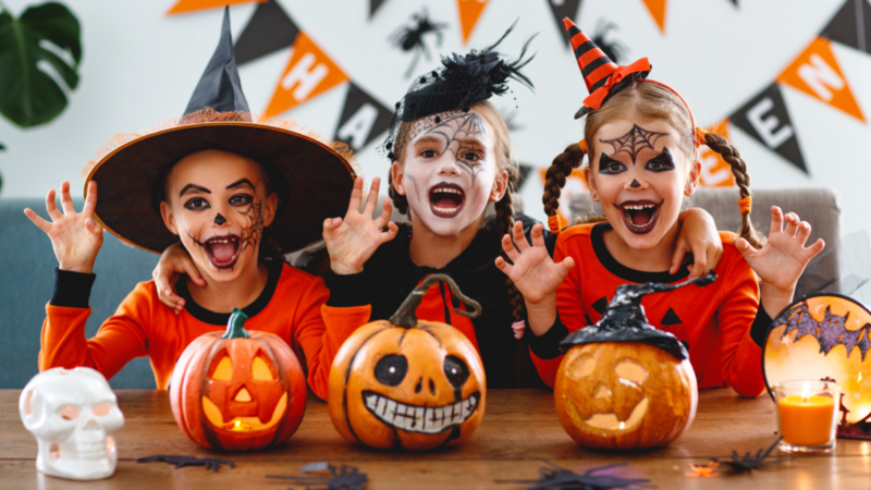 Three children in Halloween costumes sit at a table with carved pumpkins.