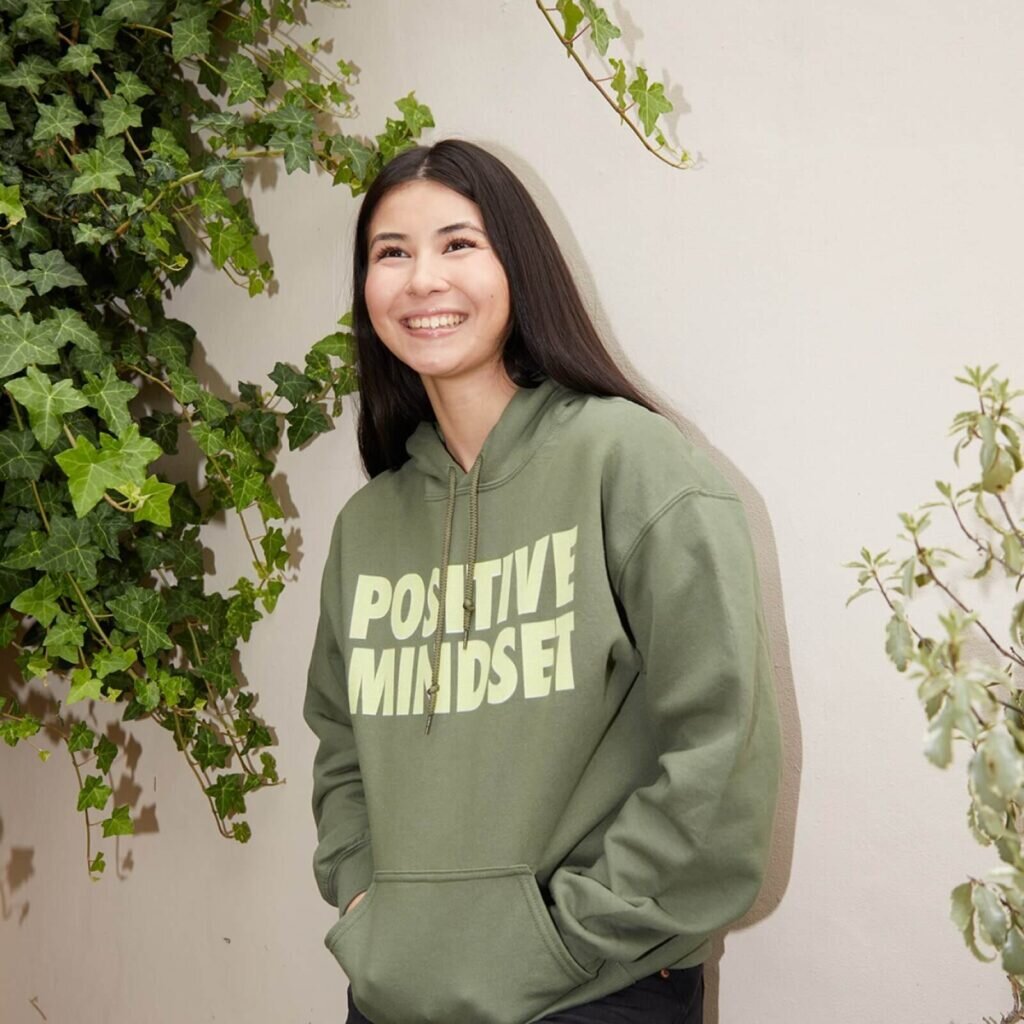 A young woman smiling and wearing a custom olive green hoodie with a text print.