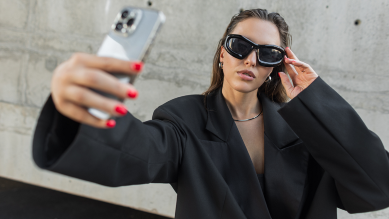 Woman in oversized black blazer and sunglasses taking a selfie with a smartphone, posing confidently against a gray concrete wall.