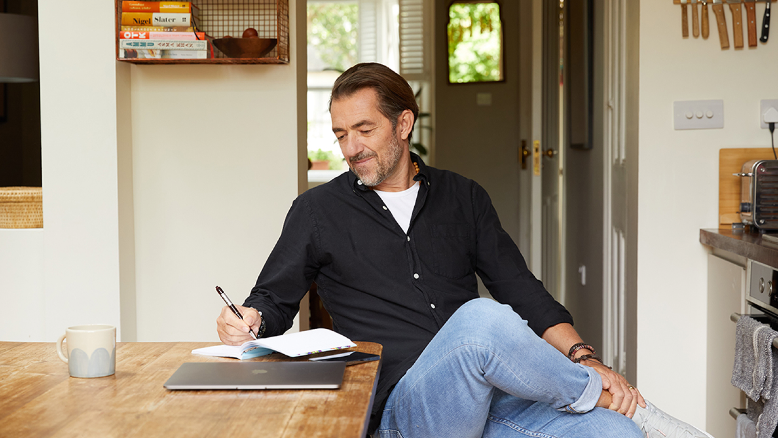 A man in a blue shirt sitting at a kitchen table, writing down something in a notebook with a laptop beside it.