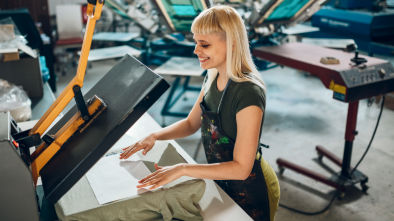 A woman with blonde hair operates a screen printing machine, smiling as she works on a green fabric.