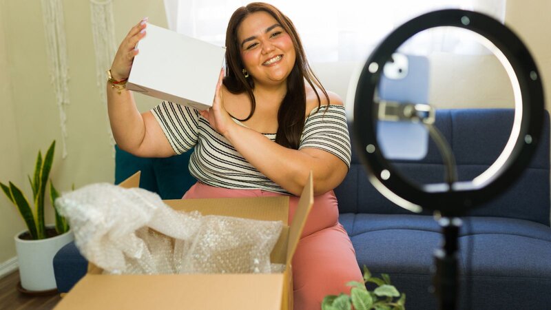 A smiling woman holding a box in front of a ring light, sitting beside an open cardboard box with packing materials, in a bright room.