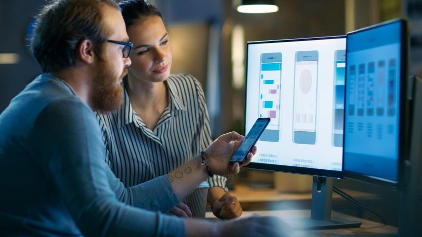 A man and a woman looking at the phone and two computer screens with designs in it.