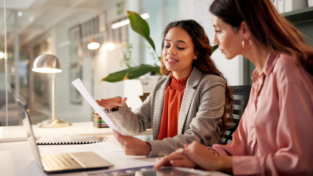 Two women seated at a desk, reviewing documents and discussing their work together.