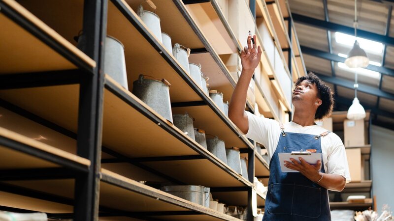 A man in overalls checking items on a high shelf in a storage room, holding a clipboard.