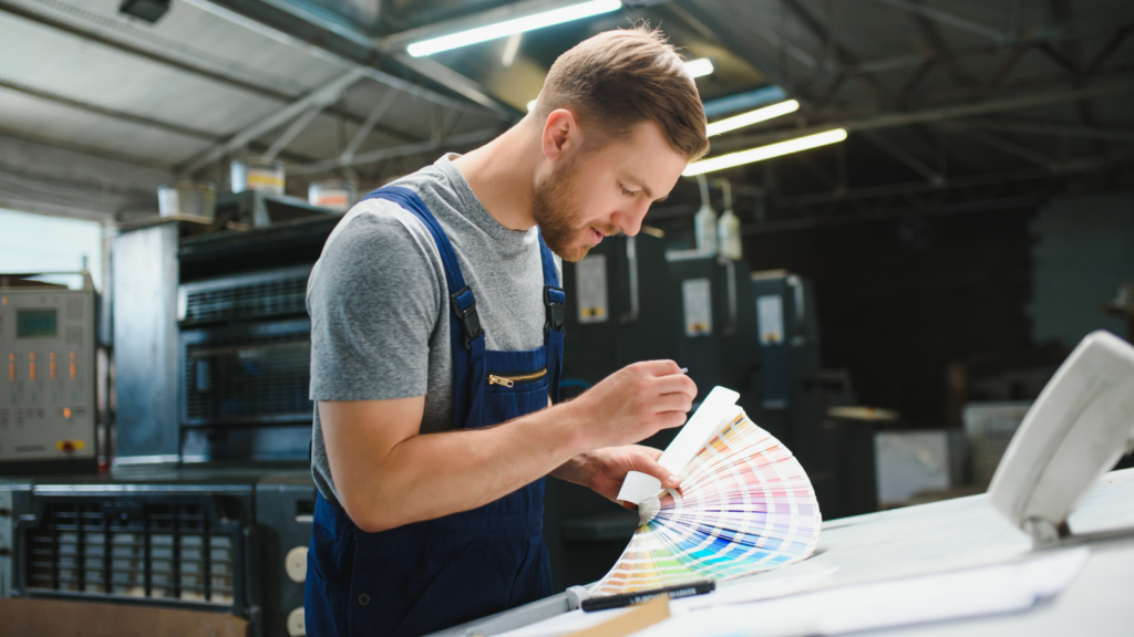 A man in an office examines a color swatch, surrounded by design materials and a computer on his desk.
