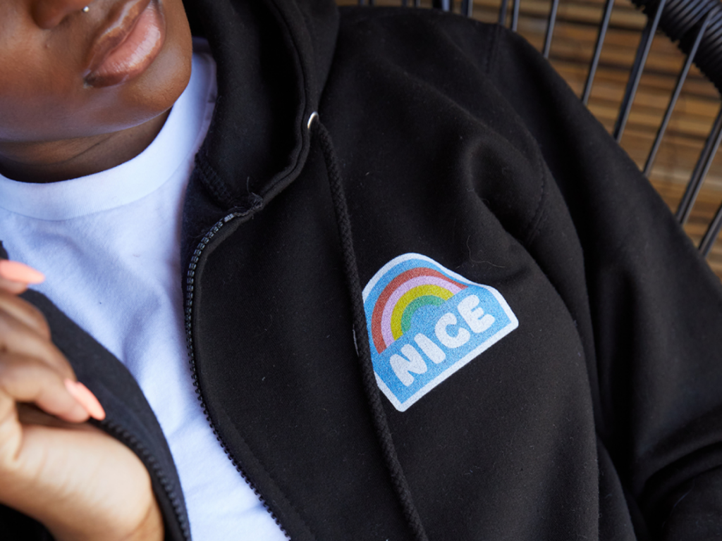 A person sitting in a chair, wearing a black hoodie with a chest print of a rainbow and the word “Nice”.