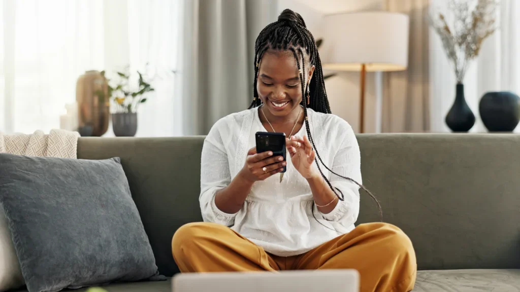 A woman with braided hair sits cross-legged on a couch, smiling at her phone. She's wearing a white top and mustard pants. The setting is cozy and relaxed.
