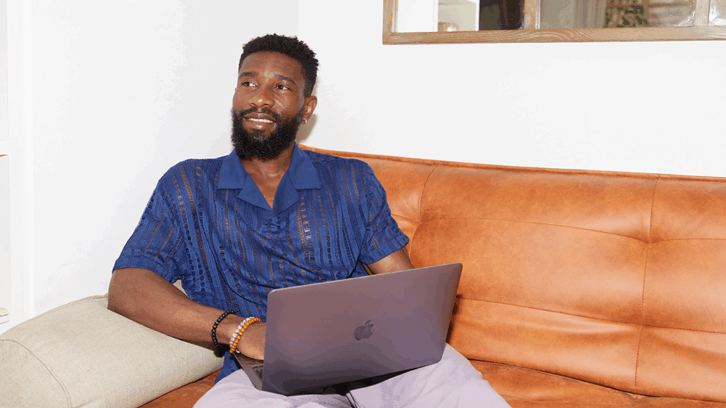 A smiling man in a blue shirt is sitting on a couch and using a laptop.